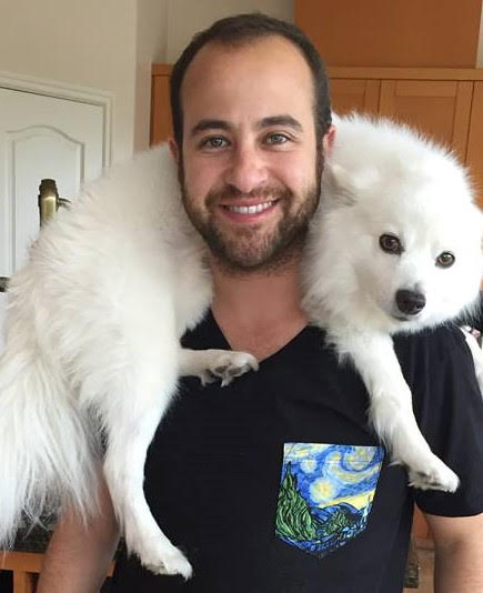 This photo contains a white passing non binary person with short brown hair & a brown beard with hazel eyes smiling.  The person is wearing a black shirt with a pocket that has Van Gogh's a Starry Night painting which has blue & white swirls with yellow swirls, and green and grey colors. With a white fluffy dog around their neck.